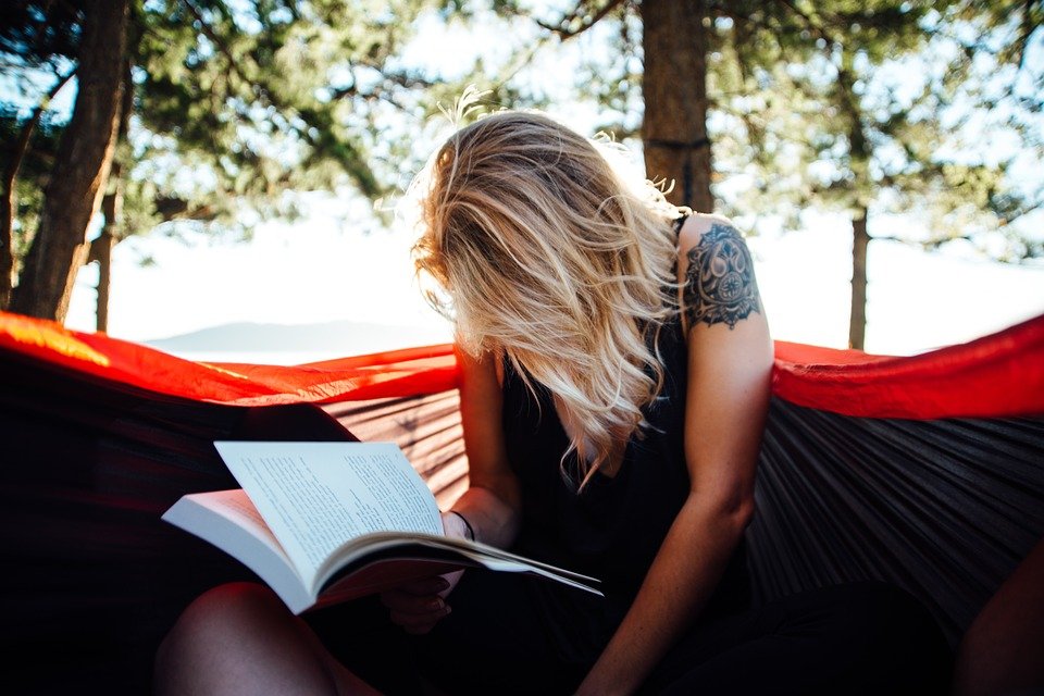 blond girl with tattoo on upper arm sitting in hammock reading and studying