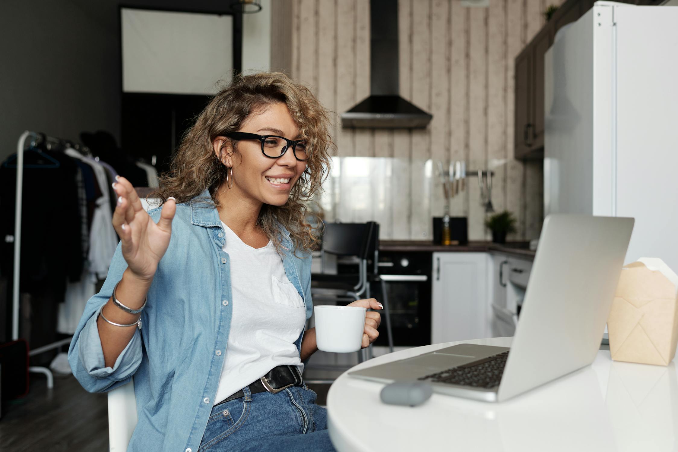 Woman in a denim shirt, holding a white cup talking to somebody via video call on laptop.