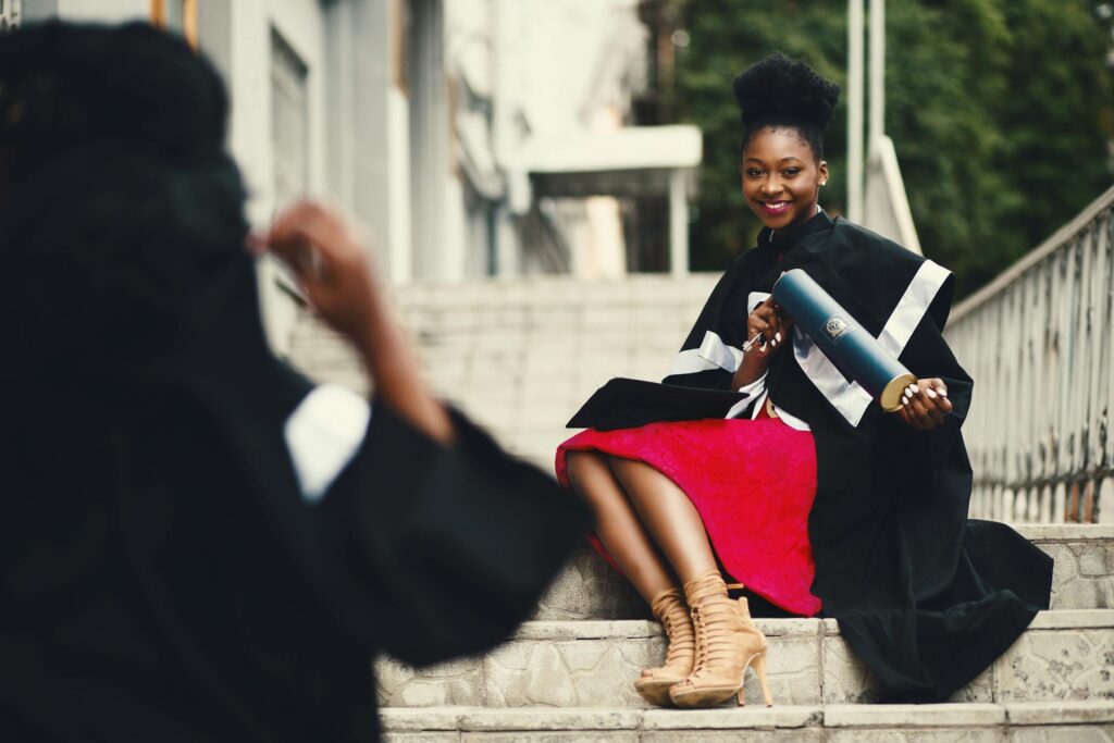 A young woman in a graduation gown and a red dress sits on outdoor steps, smiling and holding her diploma while being photographed.